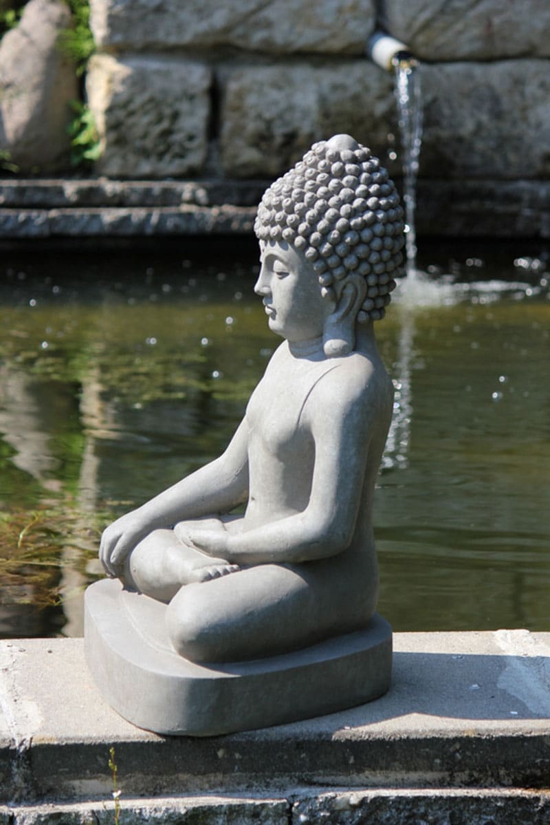 Sittande Buddha skulptur i profil vid ett vattenfall i en lummig trädgård