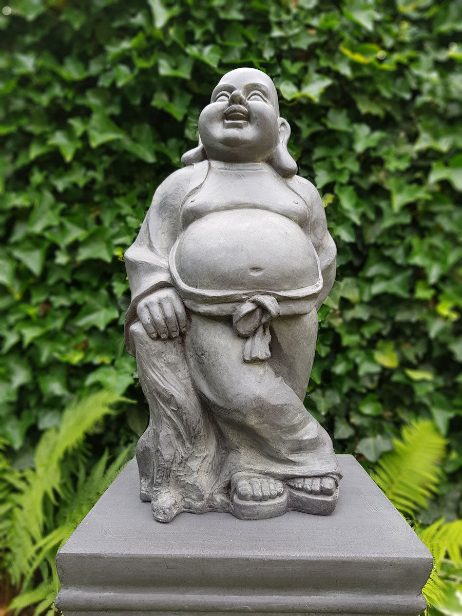 Leende Buddha trädgårdsstaty placerad på en stenpiedestal framför grön murgröna