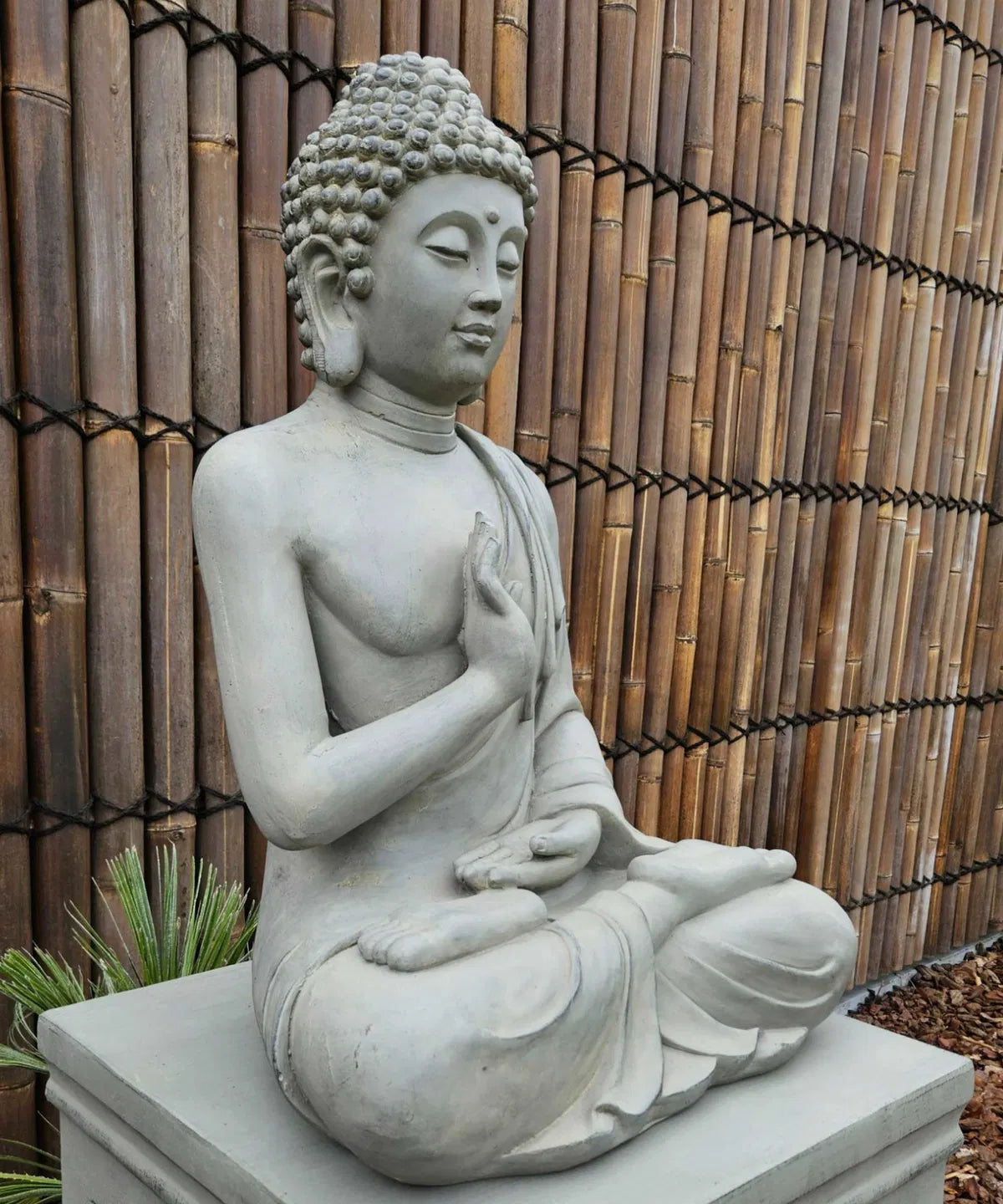 Sittande Buddha i grå finish placerad framför ett bambustaket